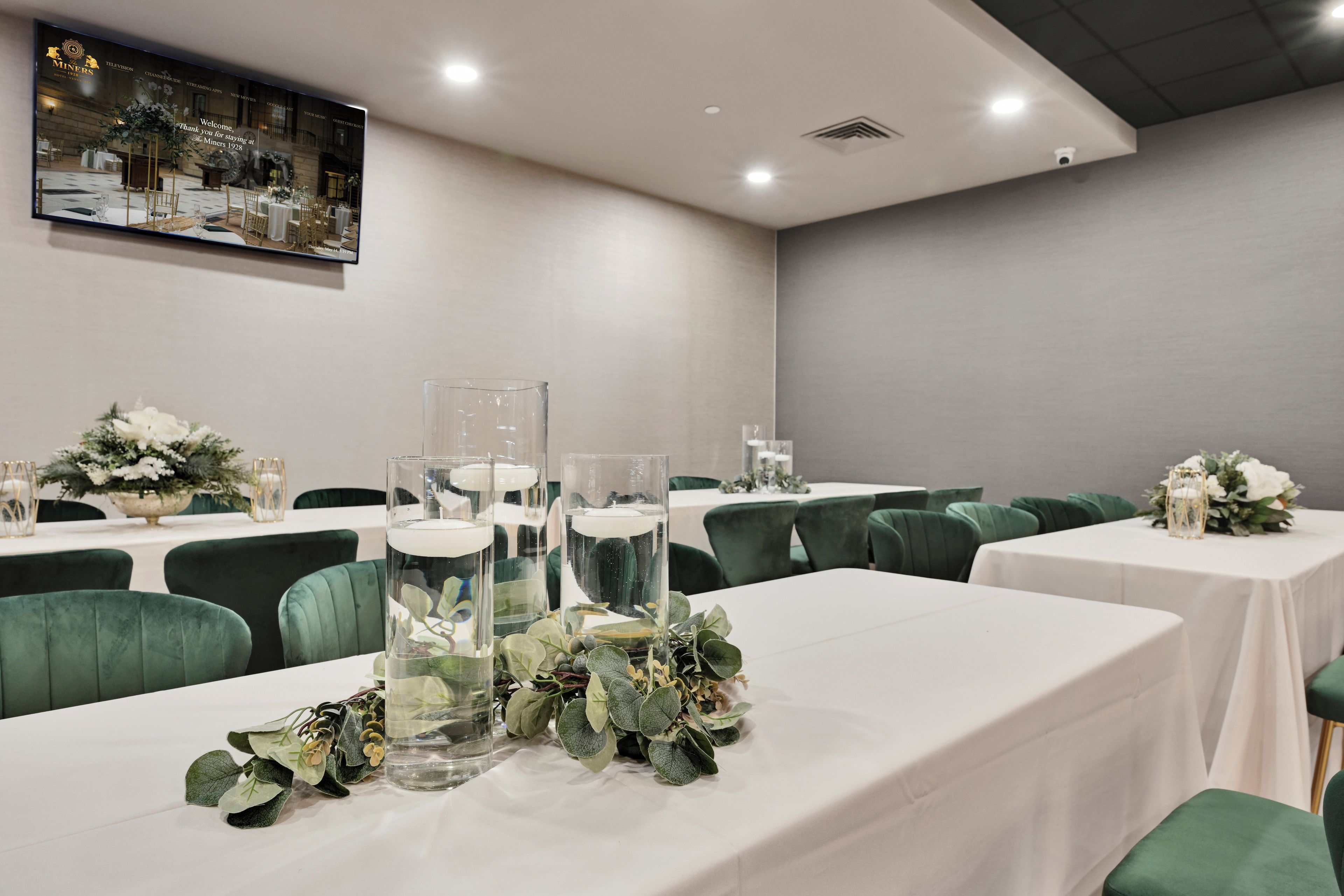 Una sala conferenze con tavoli apparecchiati per una riunione, vasi di vetro trasparente con acqua e piante verdi, e un quadro appeso al muro.