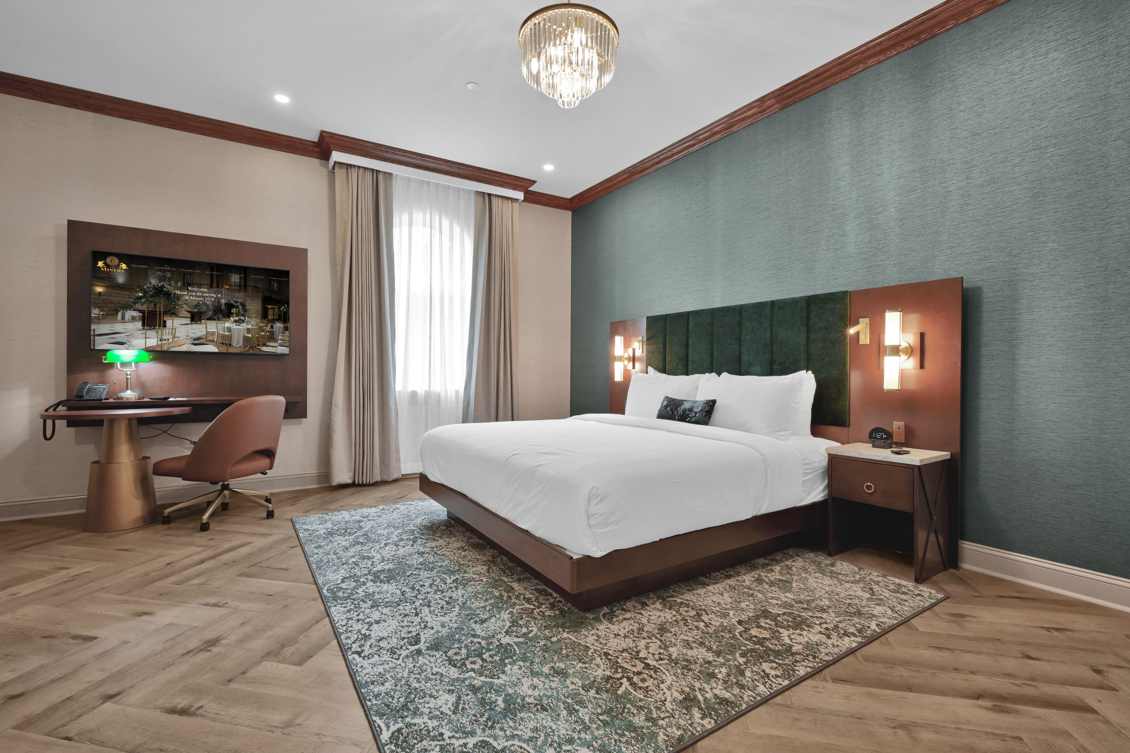A hotel room with a large bed, a desk with a computer, a chair, a desk lamp, a clock, and a wall-mounted TV.