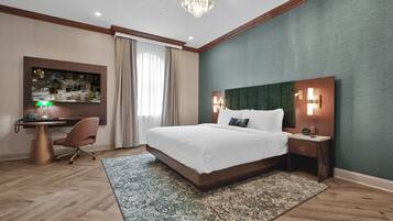 A hotel room with a large bed, a desk with a computer, a chair, a desk lamp, a clock, and a wall-mounted TV.