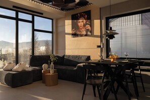 A modern living room with a sofa, dining table, and chairs, large windows with a view of mountains, and a painting on the wall. - Flowfort View Private Hottub & Sauna Cottage Lekko (Stara Kamienica)