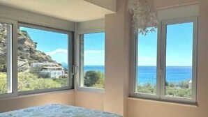 Ein Schlafzimmer mit Bett, einem kleinen Tisch und großen Fenstern mit Blick auf das Meer.