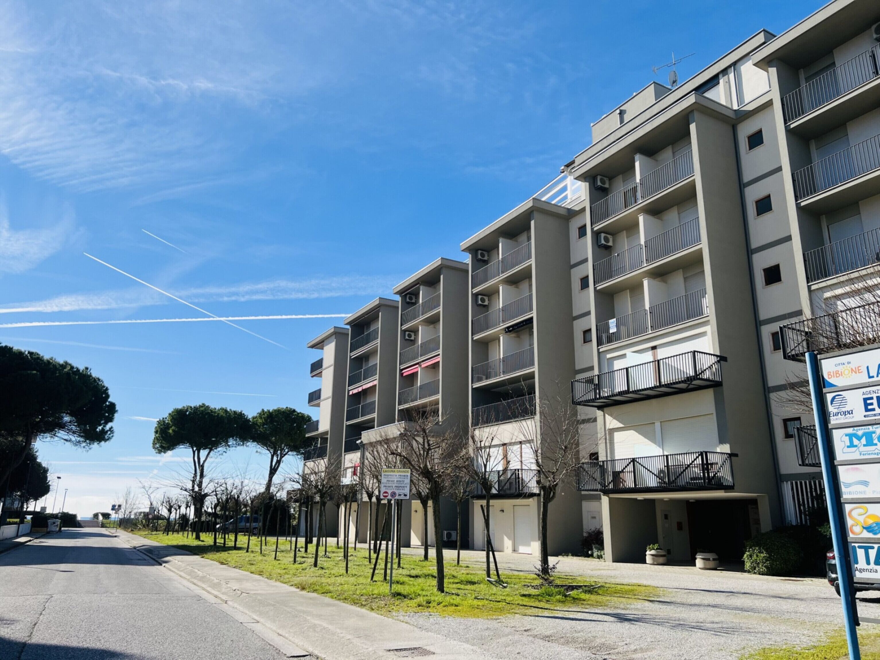 A multi-story residential building with balconies, a clear sky, and a street with trees.