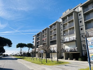 A multi-story residential building with balconies, a clear sky, and a street with trees. - Sea View Apartment in Central Bibione (San Michele al Tagliamento)