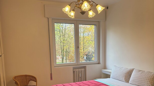 A bedroom with a wooden floor, a red bedspread, a white bed with patterned pillows, a wicker chair, and a window with a view of trees. - Spacious, light-filled apartment inside the ancient town walls, with great views (Treviso)