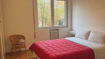 Una camera da letto con pavimento in legno, copripiumone rosso, letto bianco con cuscini a motivi, una sedia in vimini e una finestra vista alberi.