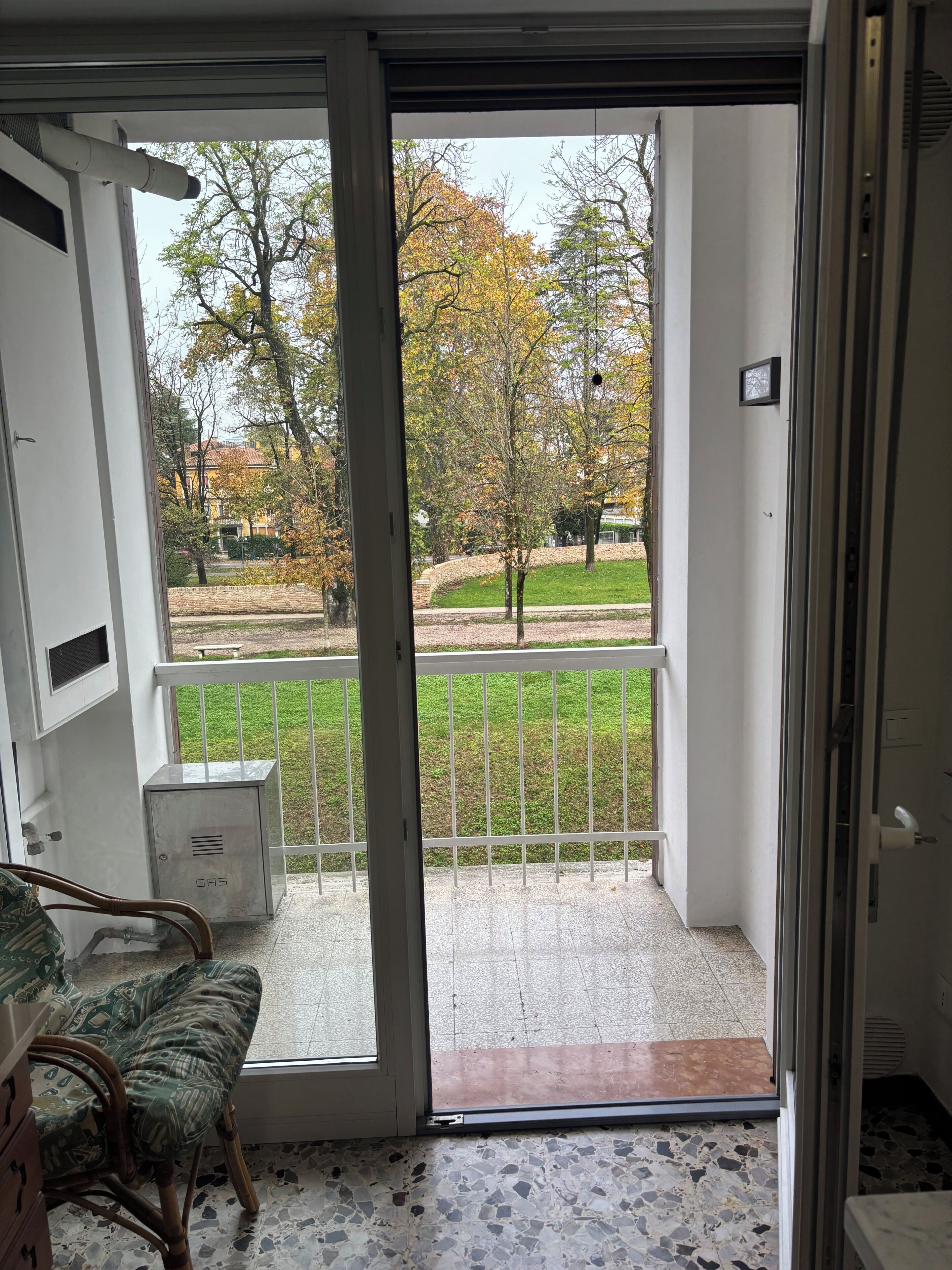A room with a sliding glass door leading to a balcony with a view of a park and trees.