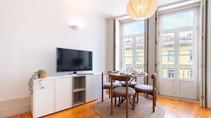 Una sala de estar moderna con un televisor de pantalla plana, una mesa de comedor con sillas, un ventanal con vistas a edificios y una lámpara colgante.