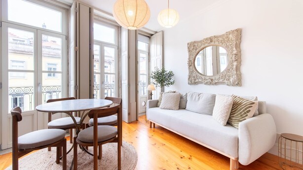 Una habitación con una mesa redonda de madera, cuatro sillas, un sofá con cojines, un espejo grande y ornamentado, y una planta en maceta.