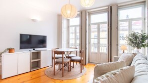 Una sala de estar moderna con una televisión de pantalla plana, una mesa de comedor con sillas, un sofá y ventanas grandes con vistas a edificios.