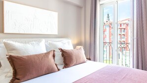 Hotellihuone, jossa on sänky, valkoiset tyynyt, ruskea heittopeitto, taulu seinällä ja parveke, jolta on näkymä rakennuksiin.