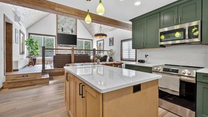 A modern kitchen with a central island, green cabinets, stainless steel appliances, and a dining area with wooden furniture. - Cabin w/ hot tub near Cana Island Lighthouse (Baileys Harbor)