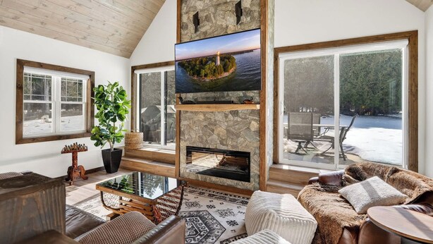 A spacious living room with a stone fireplace, large windows, and a view of a snowy landscape. - Cabin w/ hot tub near Cana Island Lighthouse (Baileys Harbor)