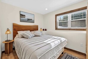 Quarto com cama, mesa de cabeceira, luminária, janela e um quadro na parede.
