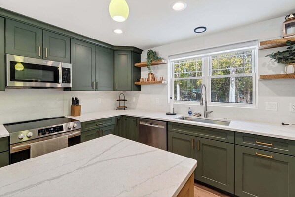 A kitchen with a large island, stainless steel appliances, green cabinets, and a window with a view of trees.