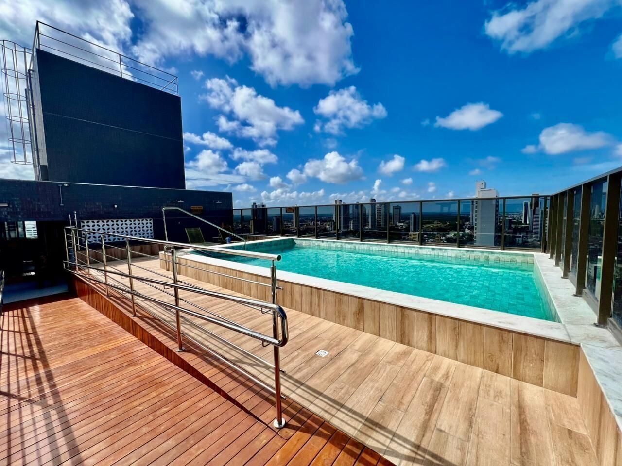 A rooftop pool with wooden deck and metal railings, surrounded by glass walls and a city view.