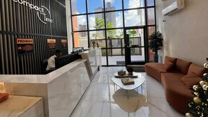 A modern reception area with a black wall, a glass partition, and a seating area with a brown sofa and a small table. - Compact Time - Miramar Hospedagens  (João Pessoa)