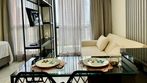 A modern living room with a glass dining table, a sofa, a black shelving unit, and a large window with curtains. - Compact Time - Miramar Hospedagens  (João Pessoa)