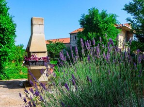 A garden with a stone structure, possibly a barbecue or oven, surrounded by lavender plants and a building with a red roof in the background. - Delux Konavle Village Apartment (Konavle)