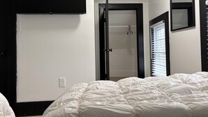 A bedroom with a bed, a door, a window with blinds, and a wall-mounted TV.