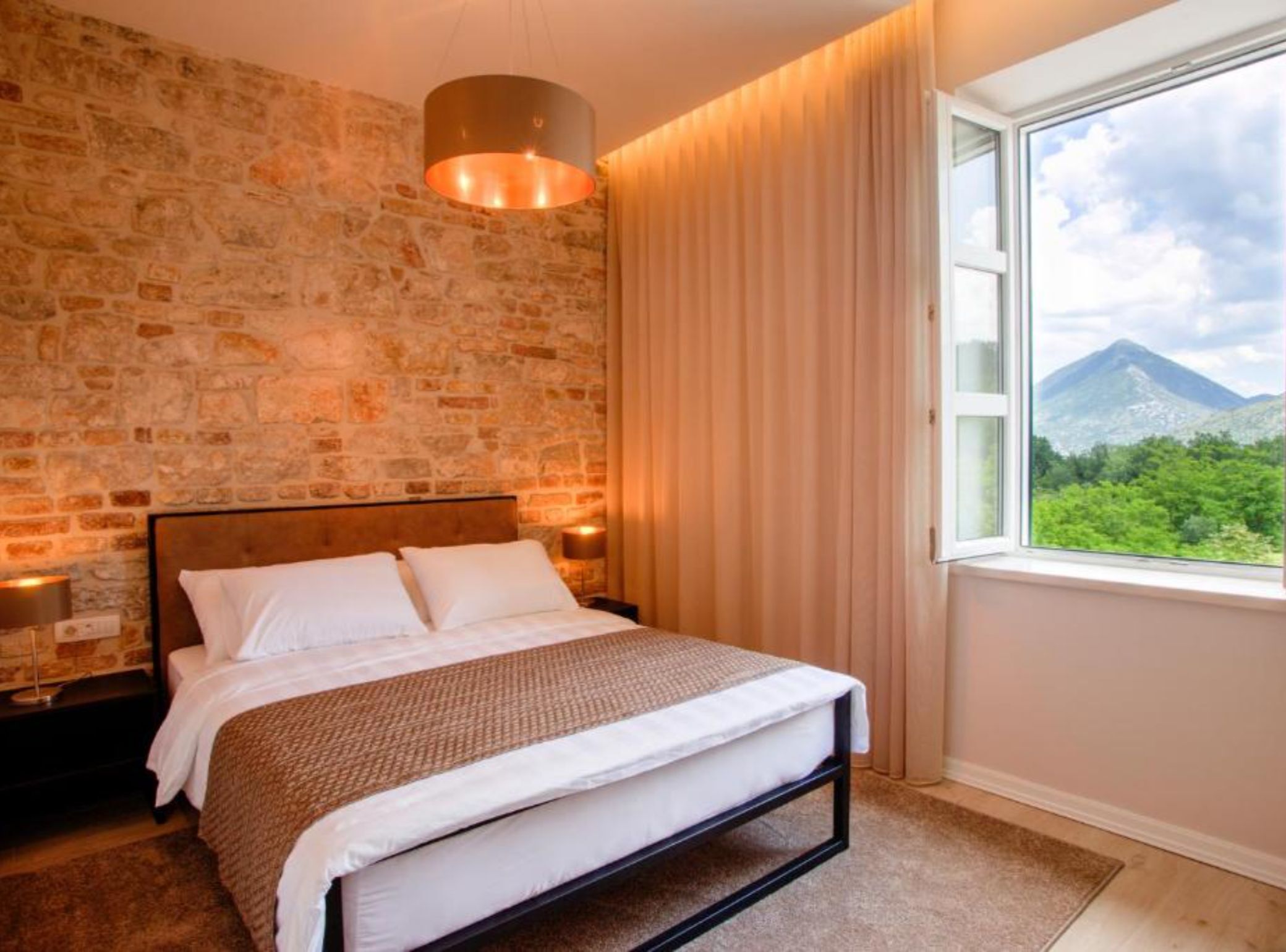 A bedroom with a stone wall, a bed with white bedding, a window with a mountain view, and a hanging light fixture.