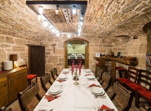 Una sala menjador de parets de pedra amb una taula llarga preparada per a menjar, cadires de fusta i una creu a la paret.