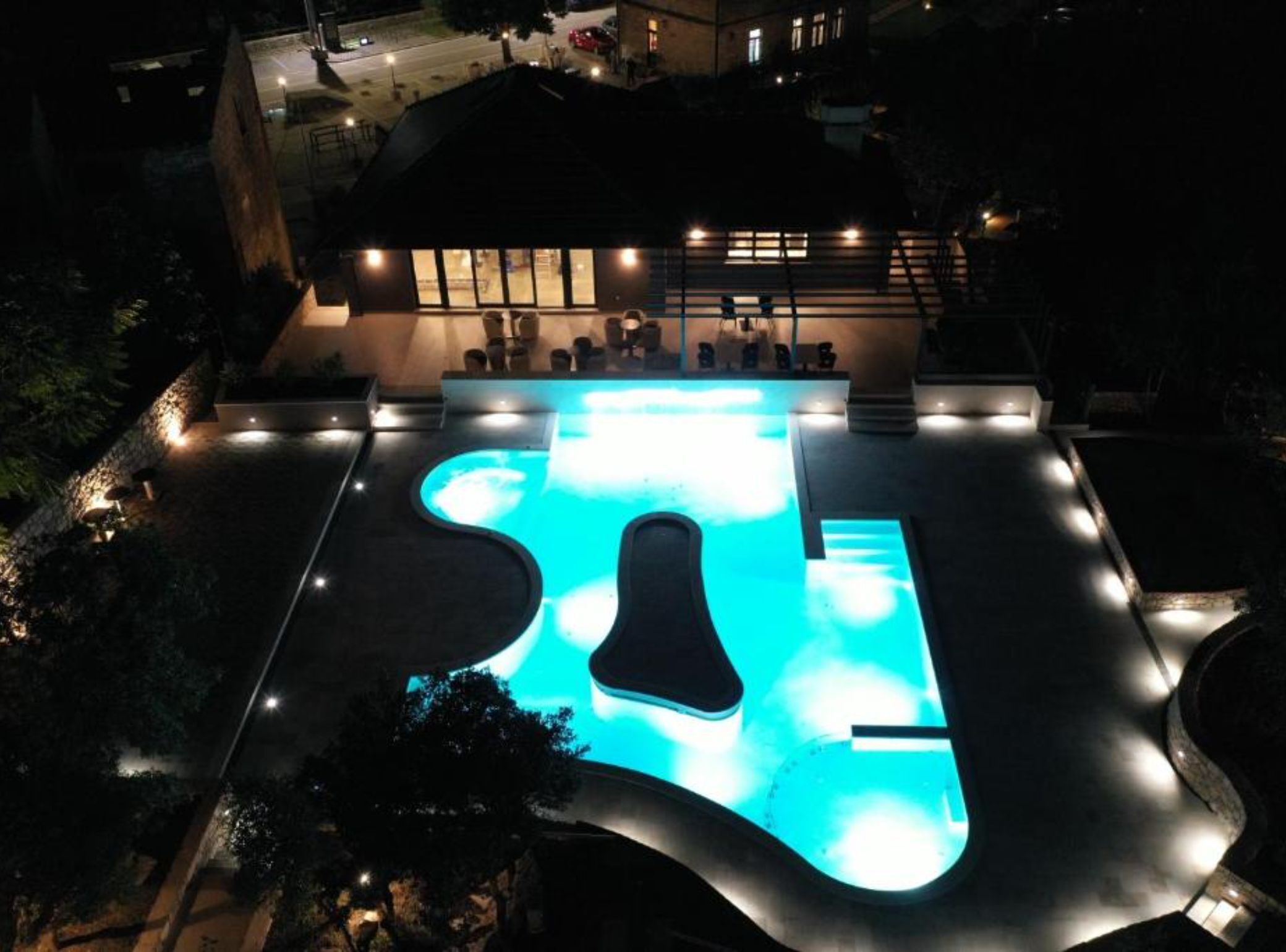 An outdoor swimming pool with a unique shape, surrounded by a well-lit deck area with seating.