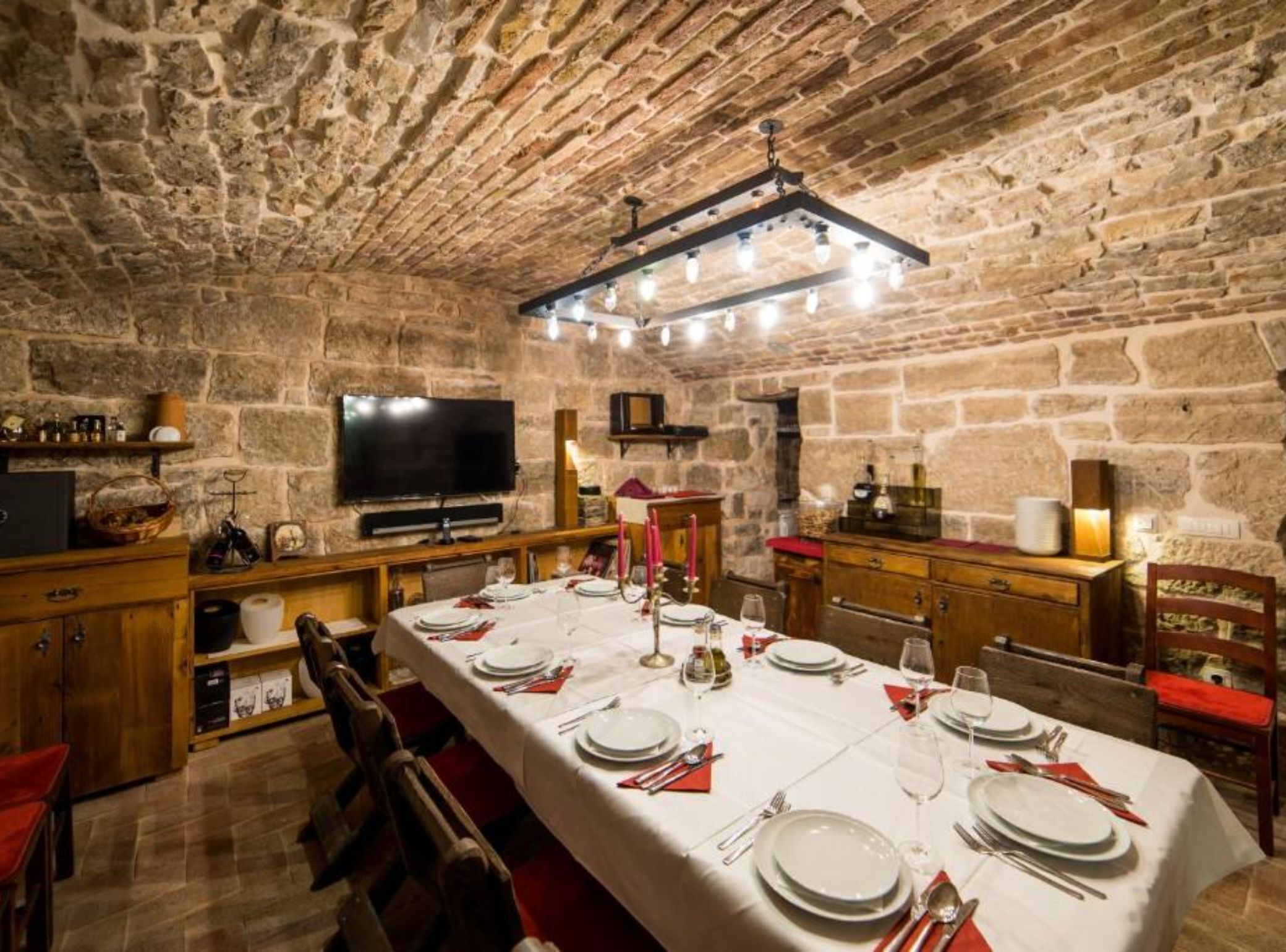 Una stanza con pareti in pietra, un lungo tavolo da pranzo apparecchiato per un pasto, mobili in legno e un televisore appeso al muro.