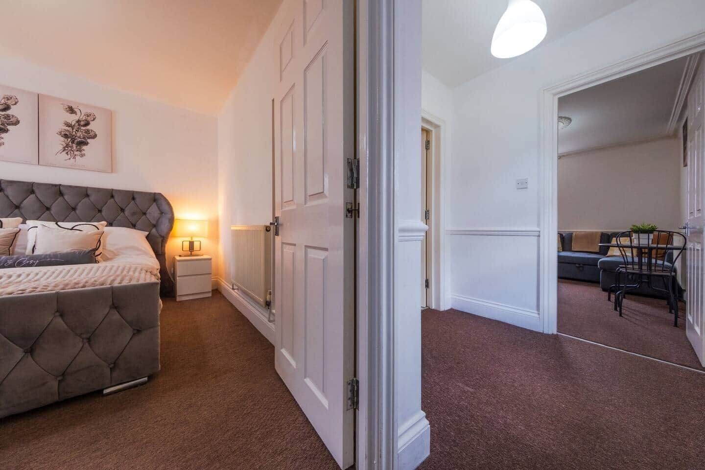 Une chambre à coucher avec une tête de lit capitonnée, une table de chevet avec une lampe, un radiateur et une porte donnant sur un espace de vie comprenant un canapé et une table à manger.