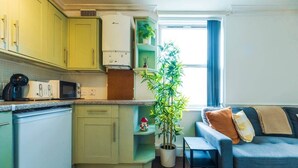 A compact kitchen with a microwave, a stove, and a sink. There is a blue sofa with cushions and a small table. A potted plant is visible near the window. - The Bristol Hidey-Hole (Bristol)