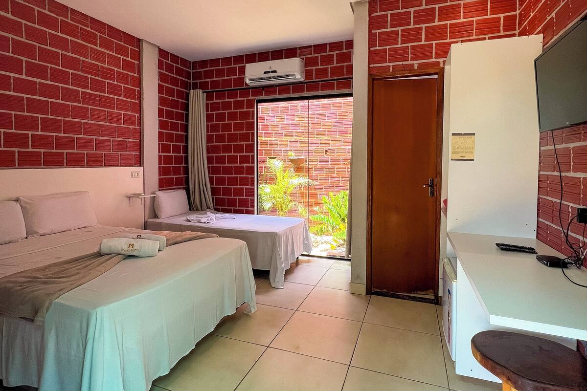 A hotel room with two beds, a desk, a TV, and a window with a view of greenery.