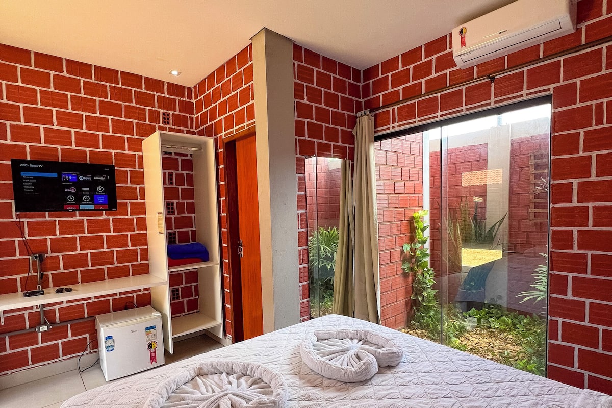 A hotel room with a brick wall, a flat-screen TV, a bed with white linens, and a sliding glass door leading to a garden.