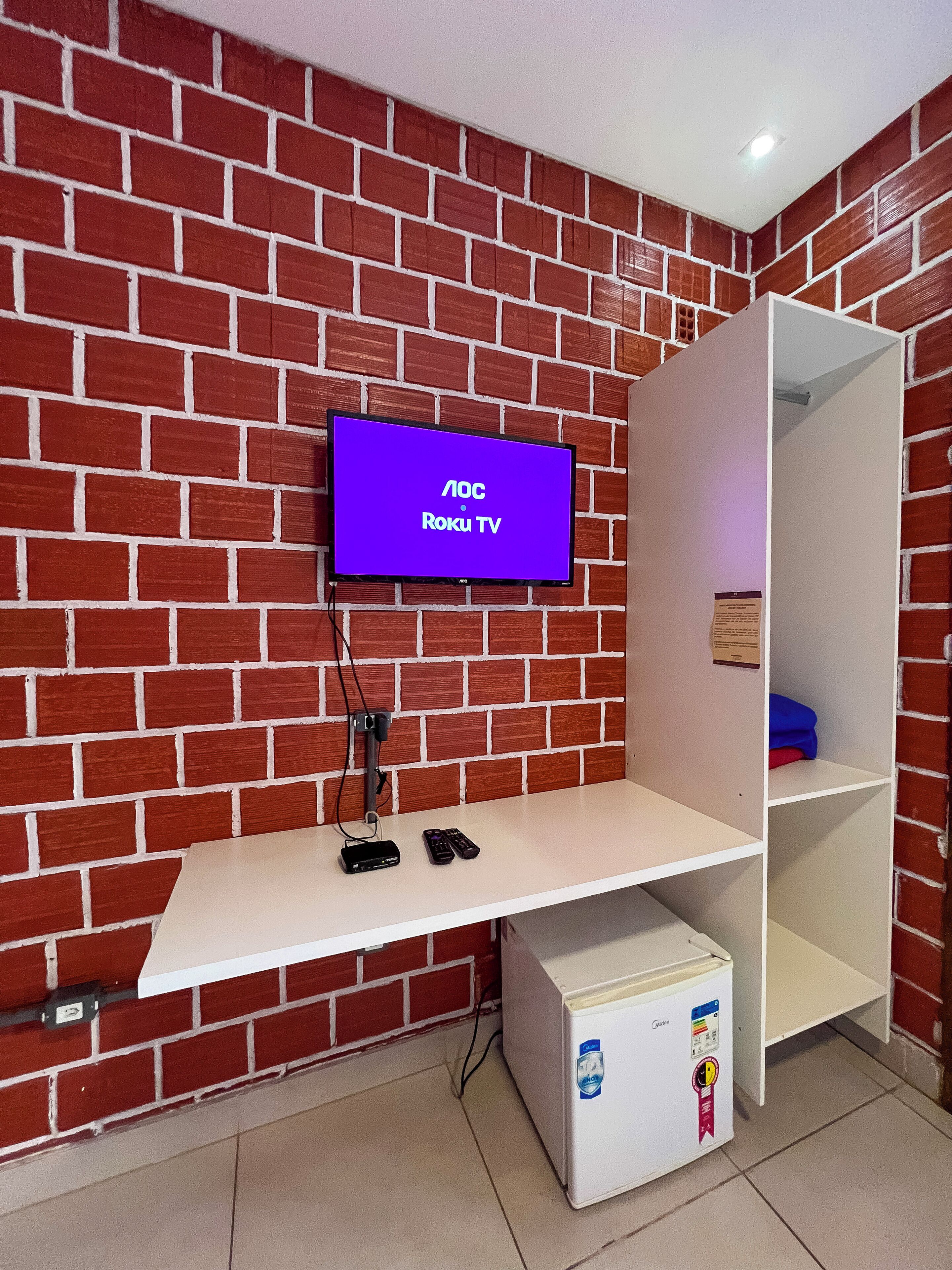 Una habitación con pared de ladrillo, un televisor montado que muestra 'LOC Roku TV', un estante blanco con dispositivos electrónicos y un mueble de almacenamiento blanco.