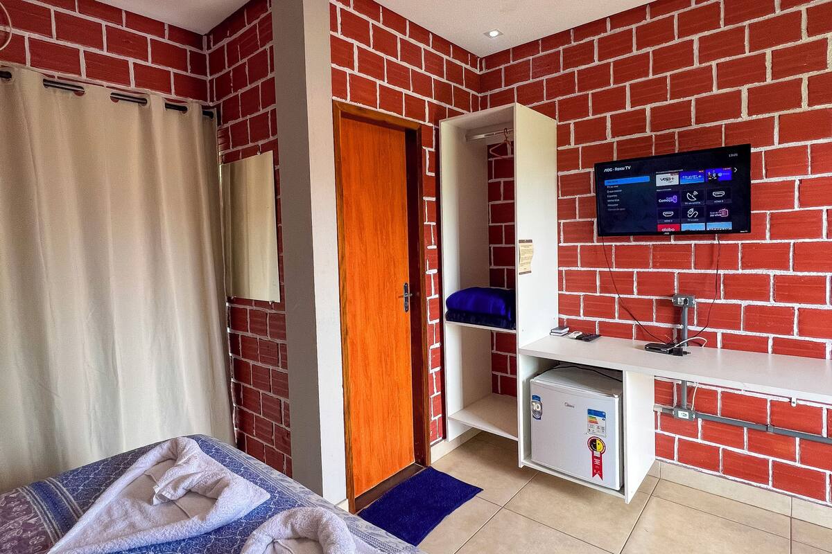 A hotel room with a brick-patterned wall, a bed with patterned bedding, a TV mounted on the wall, a wardrobe, and a small desk with a mirror.