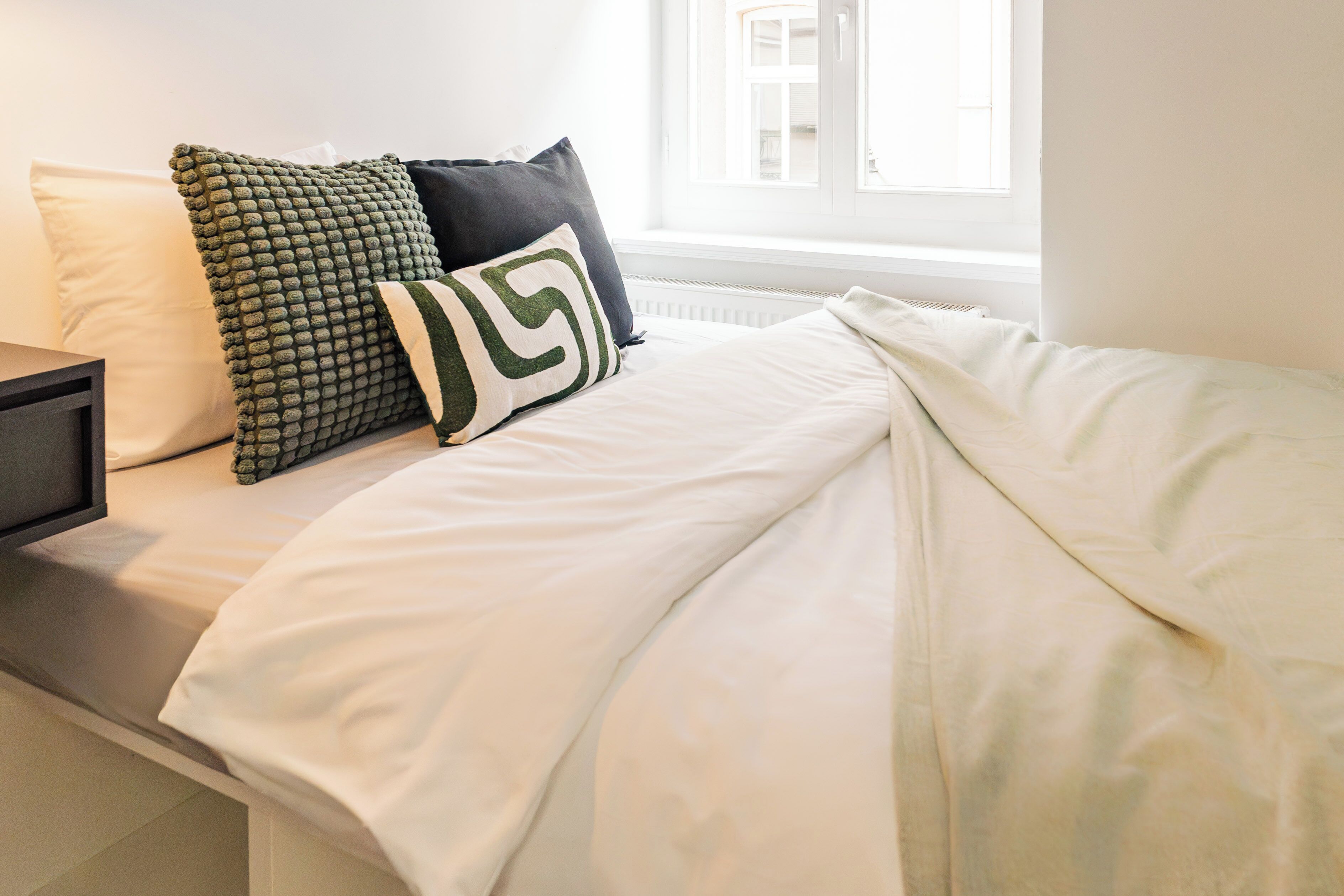 Uma cama com roupa de cama branca, um travesseiro verde e branco e um travesseiro preto.