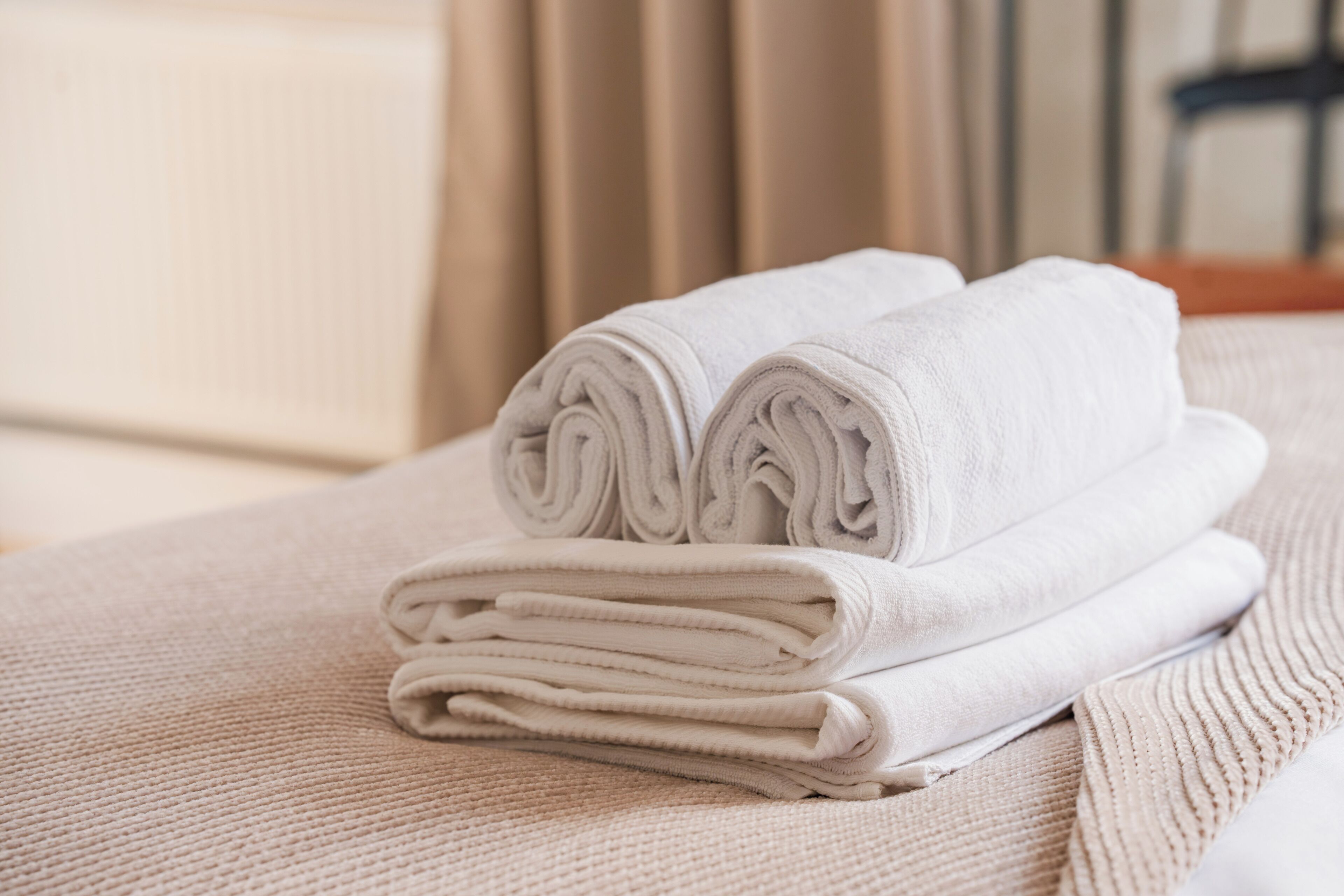 Una toalla blanca doblada con cuidado sobre una superficie beige texturizada, probablemente una cama o sofá, en una habitación con una paleta de colores neutros y persianas verticales al fondo.