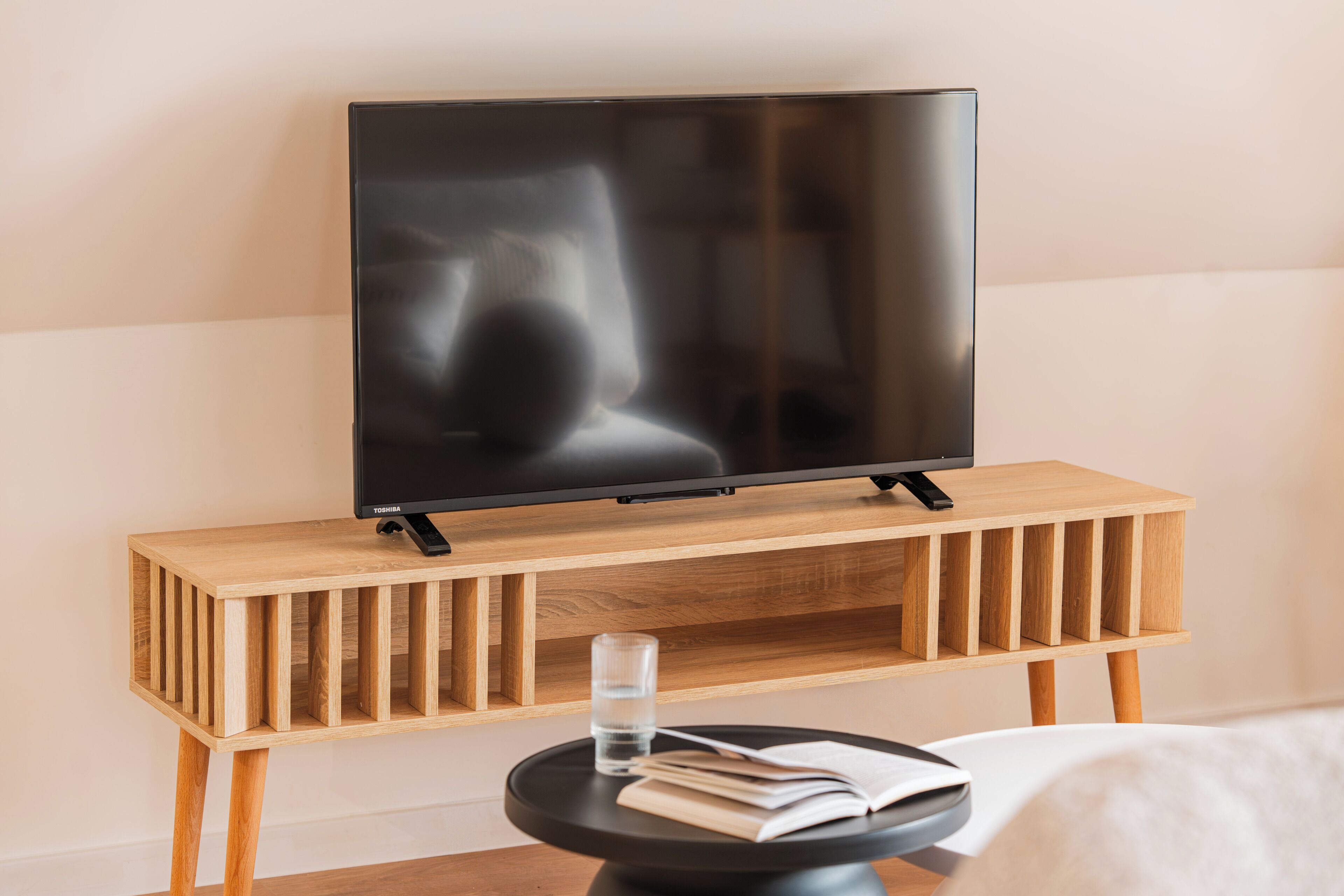 A modern room with a flat-screen TV on a wooden stand, a small round table with a book and a glass of water, and a bed in the background.