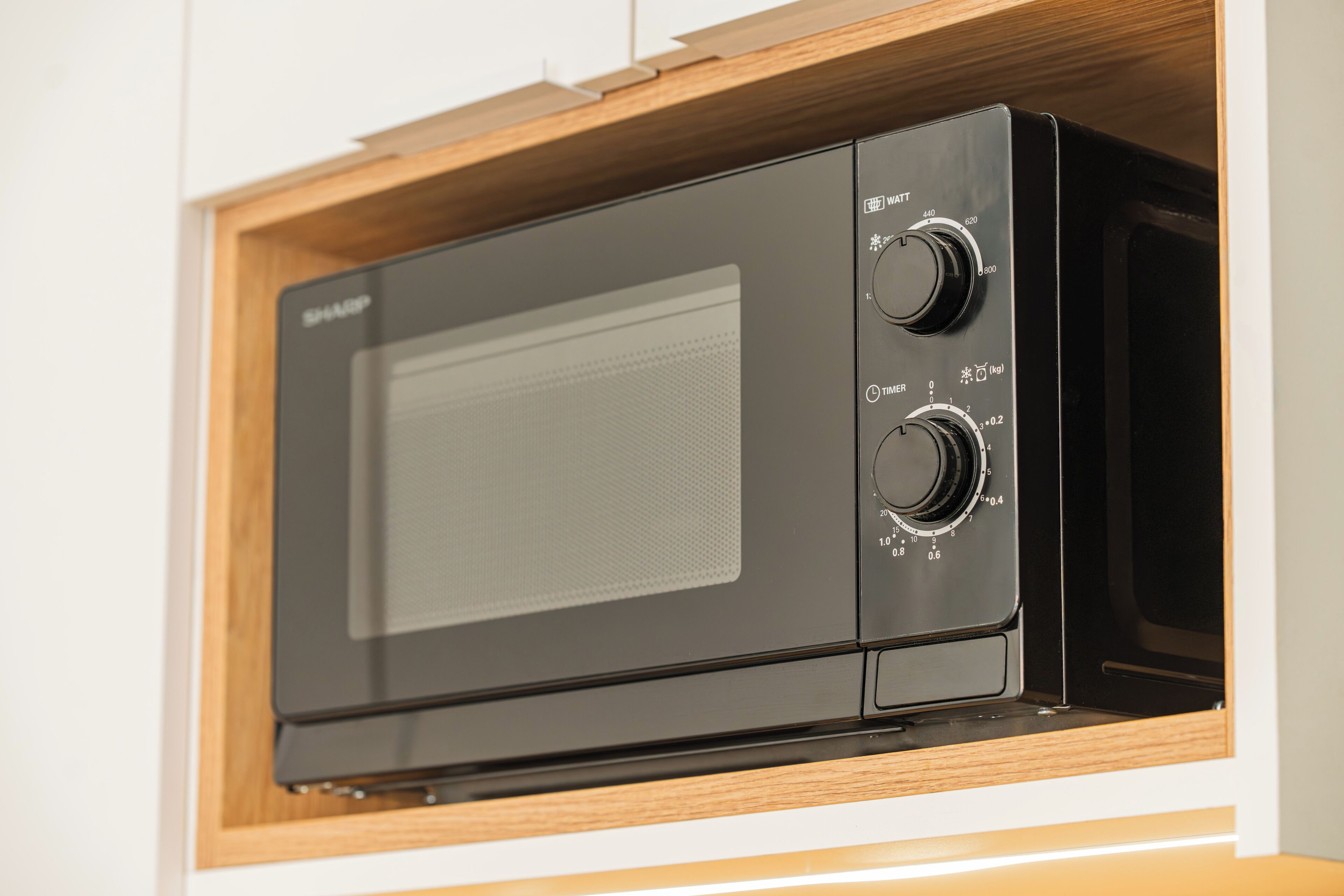 A black microwave oven with two control knobs and a digital display, housed in a wooden cabinet with a white wall.
