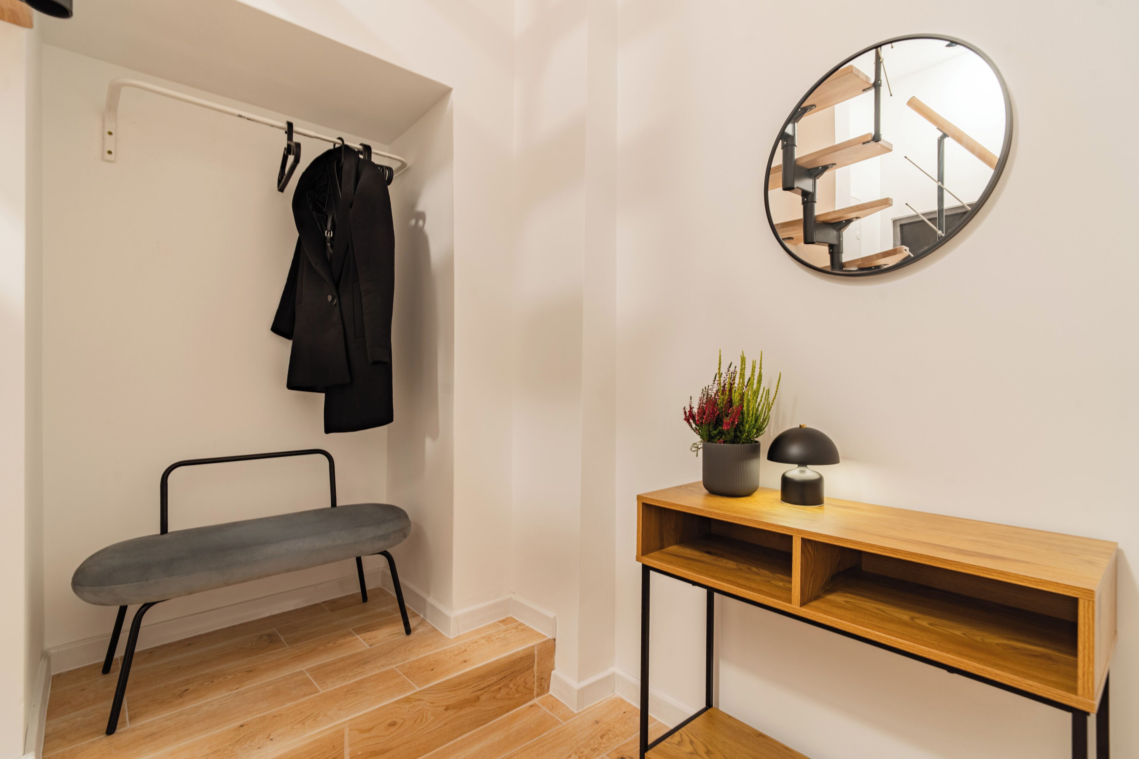 Un banc, un miroir, une plante en pot et une console en bois dans une pièce où l’on aperçoit un escalier dans le reflet du miroir.