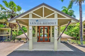 Eingangsbereich eines Resorts mit einem Schild, das 'Port Douglas Sands Resort' zeigt, umgeben von Palmen und blauem Himmel.