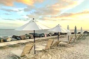 Une plage avec des chaises longues et des parasols, un rivage rocheux et un coucher de soleil en arrière-plan.