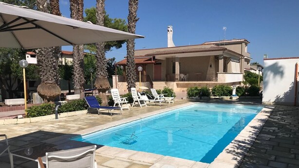 Una piscina con sillones de descanso, una mesa y un edificio con tejado de azulejos al fondo.