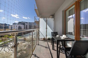 A balcony with a glass railing, a table with a teapot and cups, and a view of buildings and a park. - Spacious Apartment | 2 Bedrooms | Parking | Balcony (Kraków)