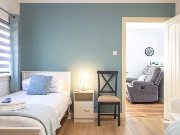 A bedroom with a bed, bedside table, chair, and a view into another room with a sofa and a clock.