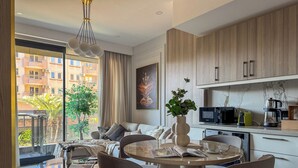 Un salon moderne comprenant un coin repas, une cuisine équipée d’un four à micro-ondes et d’une machine à café, ainsi qu’une grande fenêtre offrant une vue sur les bâtiments et la verdure.
