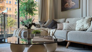 Un salon moderne avec une table basse en verre, un canapé agrémenté de coussins à motifs, une grande fenêtre donnant sur des immeubles et une œuvre d’art encadrée accrochée au mur.