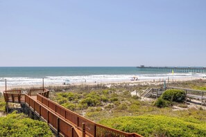 Vista de una playa con un paseo marítimo de madera, un muelle y sillas para tomar el sol.
