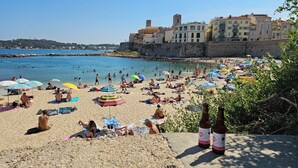 Una playa concurrida, gente tomando el sol y nadando, un muro de piedra y edificios al fondo.