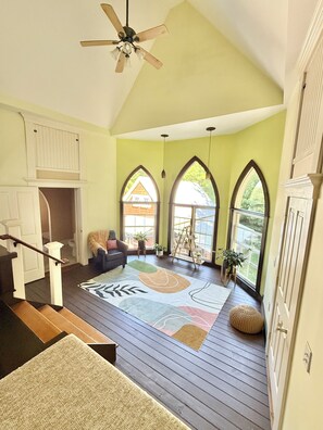 A spacious room with a high ceiling, a wooden staircase, a ceiling fan, and large windows with arched tops.