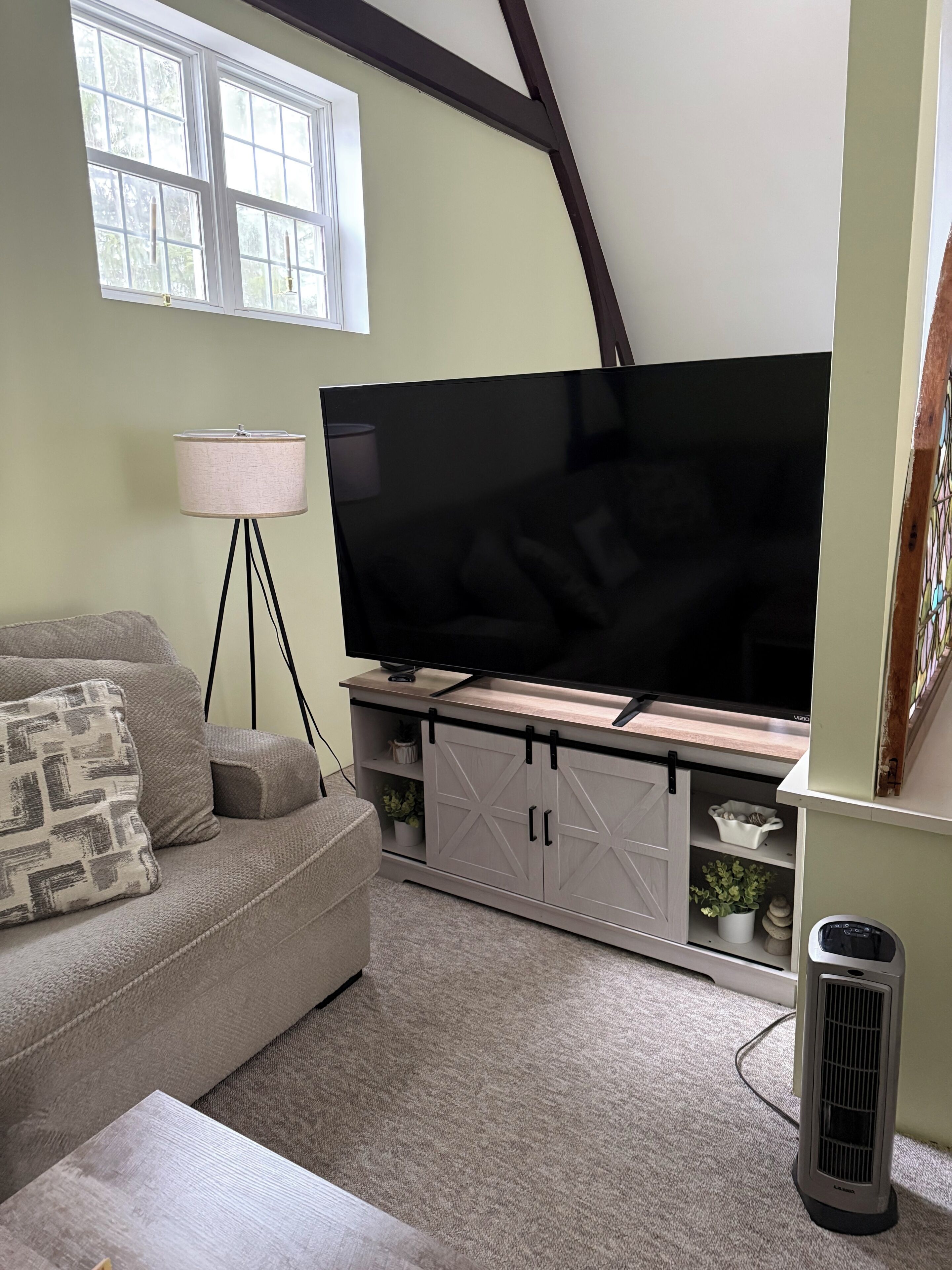 Una sala de estar con un televisor de pantalla plana sobre un soporte de madera, un sofá gris con cojines estampados, una lámpara de pie y un pequeño calentador.