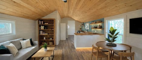 A cozy interior with a wooden ceiling, a living area with a sofa and coffee table, a kitchen with a bar, and a dining area with chairs.
